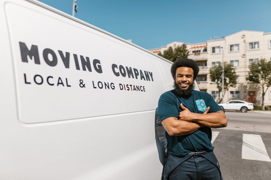 A smiling man with a detailed curly afro hairstyle and a full beard stands outdoors next to a white moving van in a city setting, representing a professional removals service. He is wearing a navy blu