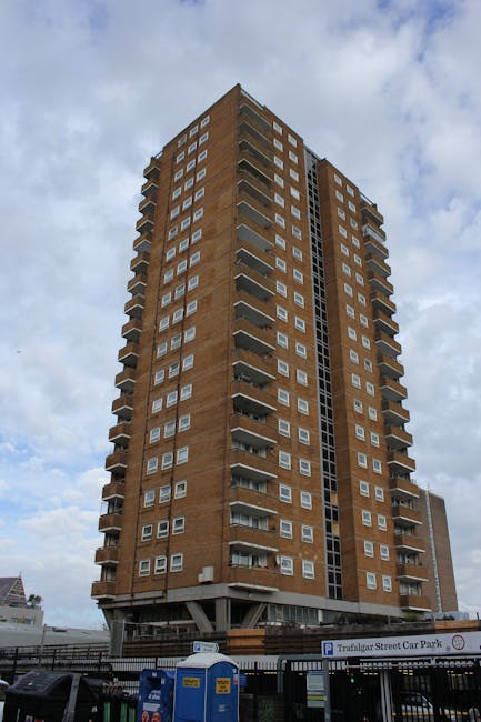 A tall, multi-storey residential building constructed from brown brick, featuring numerous small, rectangular windows and several balconies extending from the facade. The building is situated on a street with a parking area in the foreground, including a sign for Trafalgar Street Car Park, portable toilets, and a black metal fence surrounding the parking lot. The sky overhead is partly cloudy, providing natural daylight that highlights the building's external structure. This scene depicts an urban environment typical for a house or flat relocation context, where furniture and boxes might be moved to or from such flats. The image is relevant for highlighting residential removals, packing and moving activities, and the logistical considerations involved in transporting household items from high-rise flats, as offered by companies such as Man With a Van Brixton.
