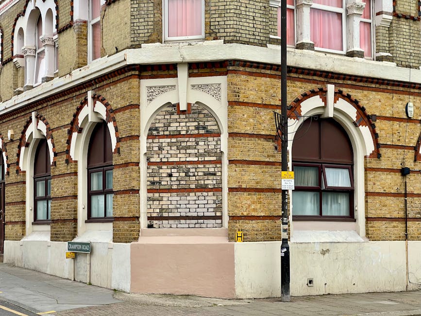 Photograph of a multi-storey brick building on a residential street, featuring large arched windows with dark frames and pink curtains inside. The building has detailed architectural elements, including decorative white trim and a mixture of light and dark bricks. A black lamppost with a yellow and white bus stop sign and a small yellow ‘H’ sign are positioned on the sidewalk in front of the property. The ground-level facade shows a pale pink section, possibly a small step or platform, leading to a central area boarded up with bricks, indicating possible ongoing repairs or alterations. The street sign ‘Crawton Road’ is mounted on a short post nearby. The overcast natural lighting creates a neutral environment typical of urban areas engaged in home relocation and furniture transport activities. This scene captures the exterior environment where professional removals, such as those by [COMPANY_NAME], might prepare for a house move involving loading furniture and packaging materials, emphasizing the importance of organized loadings during home relocation processes.