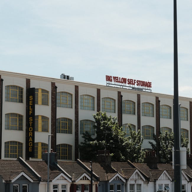 A tall, multi-storey residential building constructed from brown brick, featuring numerous small, rectangular windows and several balconies extending from the facade. The building is situated on a street with a parking area in the foreground, including a sign for Trafalgar Street Car Park, portable toilets, and a black metal fence surrounding the parking lot. The sky overhead is partly cloudy, providing natural daylight that highlights the building's external structure. This scene depicts an urban environment typical for a house or flat relocation context, where furniture and boxes might be moved to or from such flats. The image is relevant for highlighting residential removals, packing and moving activities, and the logistical considerations involved in transporting household items from high-rise flats, as offered by companies such as Man With a Van Brixton.