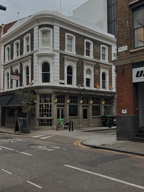 A multi-story building located on a city street corner with a grey and white facade, featuring large ground-floor windows and decorative architectural details. Outside, there is a bicycle with a bright green cover parked against the building, along with black bollards and a small chalkboard sign on the sidewalk. The street has road markings including bicycle lanes, with a double yellow line along the curb. The environment is urban with other buildings nearby, and the setting appears suitable for home relocation or furniture transport services, as provided by Man With a Van Brixton. No people or moving equipment are visible in this image.