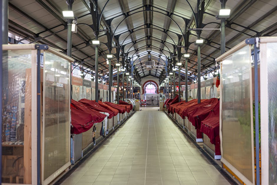 Inside a spacious market hall with a high, arched ceiling supported by dark metal beams and evenly spaced rectangular lighting fixtures. The corridor is lined with wooden display stalls covered with transparent plastic panels and red fabric drapes. At the far end of the hall, there is an open doorway allowing natural light to illuminate the interior. The floor is tiled with light-colored tiles. To the right, a partially visible mobile storage unit or trolley with a metallic frame is positioned along the aisle, and some packing boxes or containers are visible on the stalls, indicating a setting related to house removals or moving logistics. The overall scene depicts an organized space, suitable for loading or unloading items during a home relocation, aligning with the services offered by Man With a Van Brixton for same-day removals in Brixton Market and Electric Avenue.